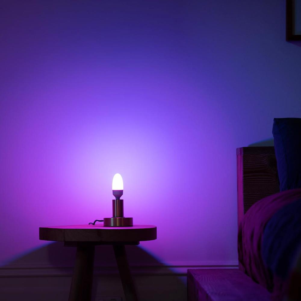 A LIFX smart candle shaped, E14 bulb in a lamp glowing a violet blue colour on to a background wall