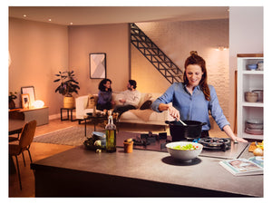 3 people in an open plan home.  2 people on a sofa in the background and 1 person cooking food in the foreground