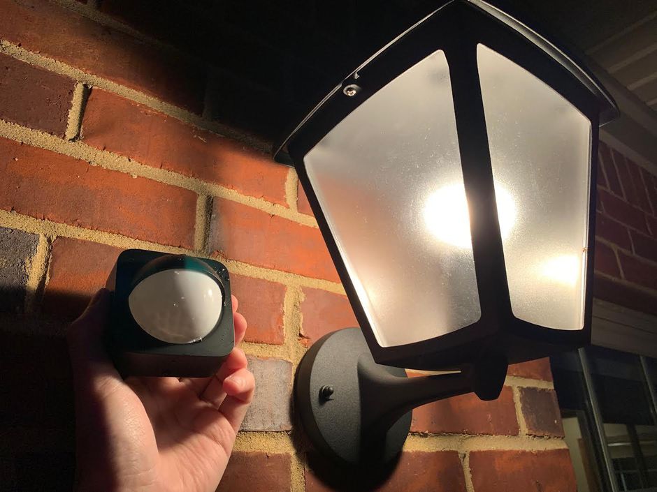 A brick wall with a lantern mounted to it and a hand holding a philips hue smart motion sensor next to it