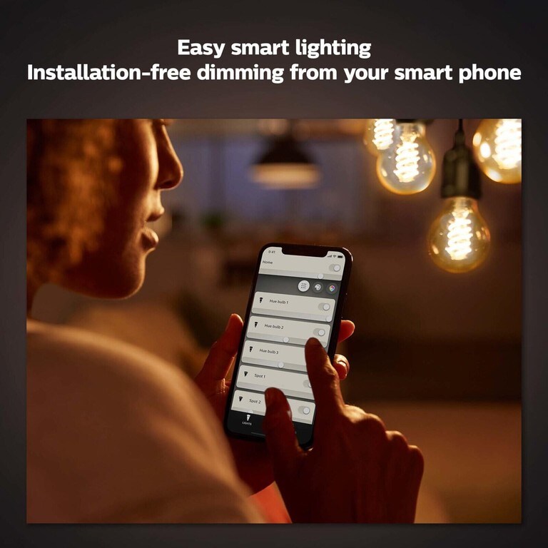 A person using a mobile phone with the philips hue app to control the philips hue filament smart light bulbs dangling in the background